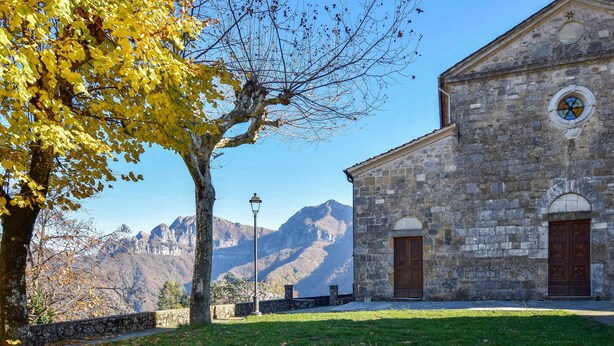A stone building with a clock and two doors, surrounded by a grassy area and a tree with yellow leaves. - Gorgeous home in Farnocchia with WiFi (Farnocchia)