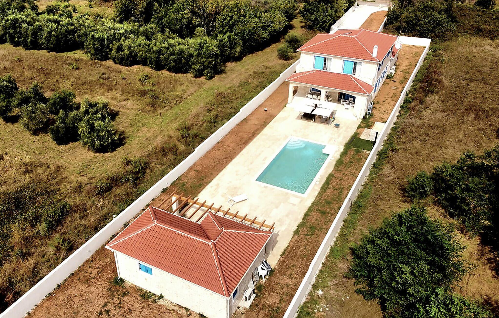 Uma casa de dois andares com piscina, cercada por vegetação e um acesso pavimentado.