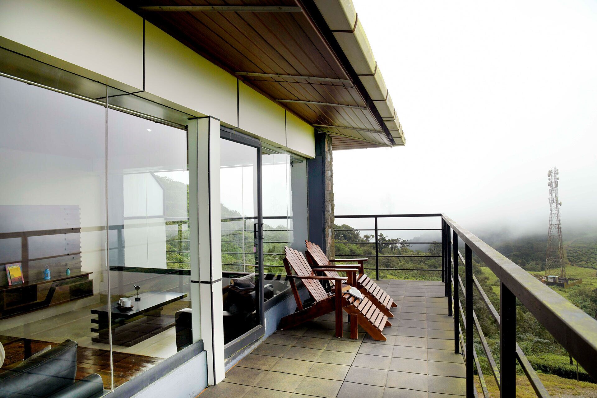 A balcony with wooden chairs, a table, and a glass wall overlooking a landscape with a power line tower.