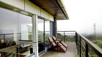 A balcony with wooden chairs, a table, and a glass wall overlooking a landscape with a power line tower.