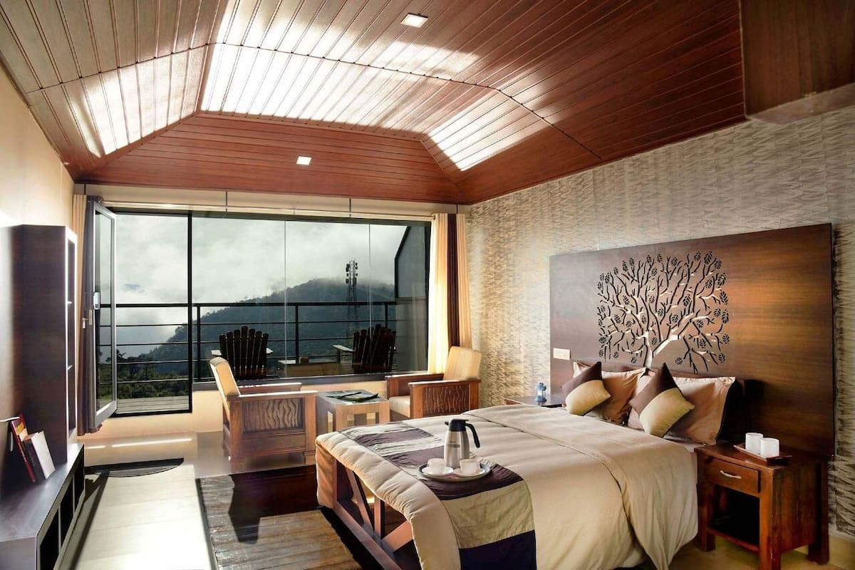 A modern hotel room with a large bed, a wooden ceiling, a stone wall, and a large window offering a mountain view.