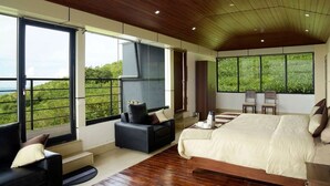 A modern bedroom with a large bed, a sofa, and a TV. There are large windows offering a view of greenery. - Ibex Resorts, Valparai (Tale Rock) (Valparai)