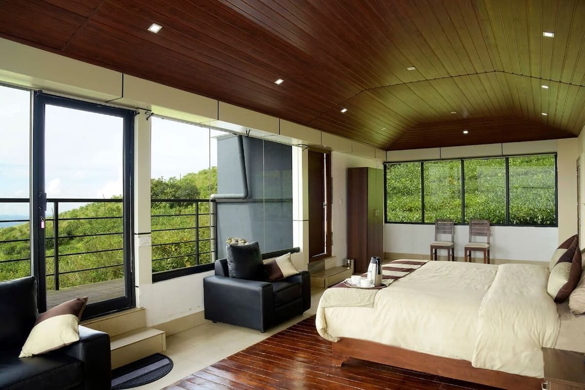 A modern bedroom with a large bed, a sofa, and a TV. There are large windows offering a view of greenery.