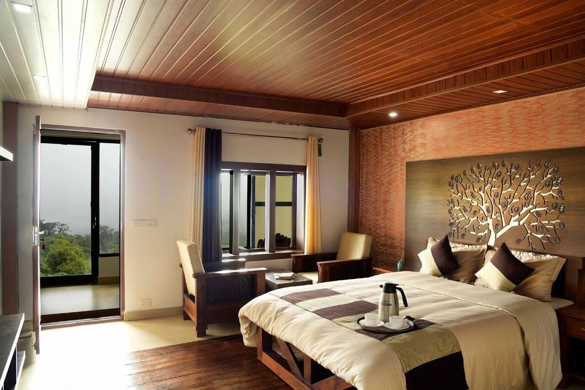 A modern hotel room with a large bed, a desk, and a chair. There is a window with curtains, a view of greenery outside, and a tree mural on the wall.
