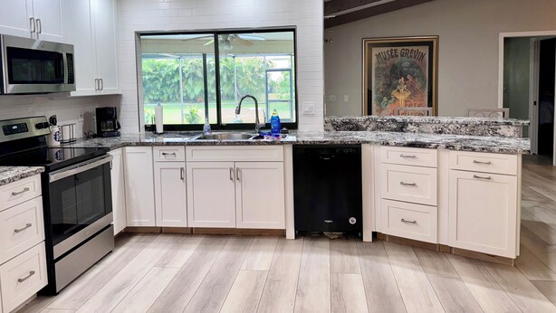 A modern kitchen with white cabinets, a granite countertop, a built-in microwave, a dishwasher, and a large window offering a view of greenery. - CHARMING! 3-bedroom house with Pool, WiFi, AC in marvelous Fort Myers near beach (Fort Myers)
