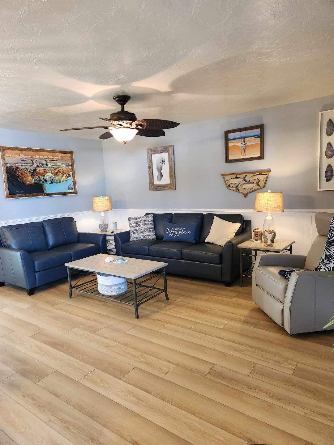 Una sala de estar con un sofá azul, un sillón blanco y una mesa de centro. Se observan obras de arte en las paredes y un ventilador de techo.