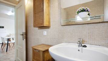 Une salle de bain avec une grande baignoire blanche, des meubles en bois, une étagère en verre ornée d’une plante en pot et un mur recouvert de carreaux.