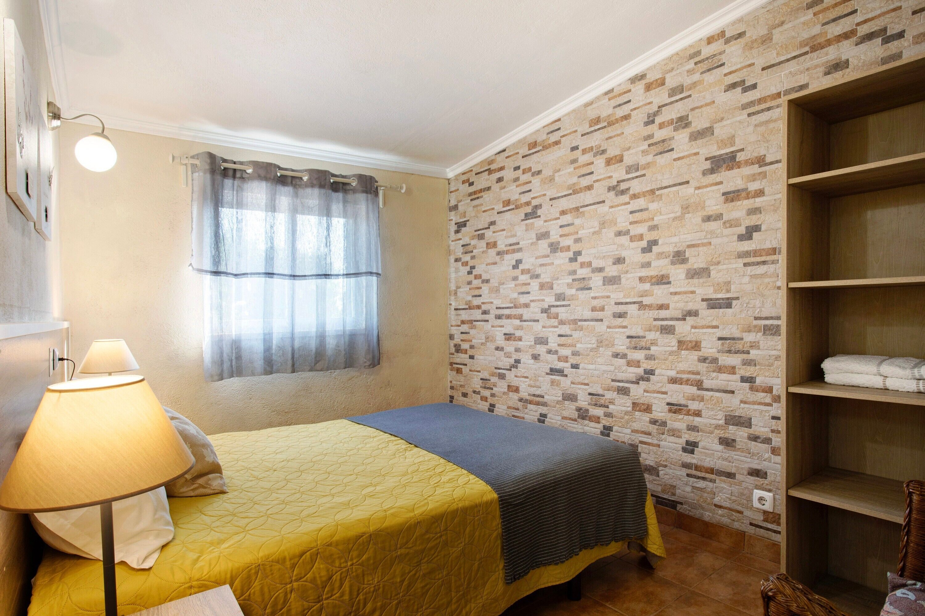 A bedroom with a bed, a lamp, a window with curtains, a brick-patterned wall, and a wooden shelf.