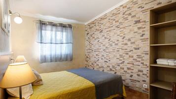 A bedroom with a bed, a lamp, a window with curtains, a brick-patterned wall, and a wooden shelf.