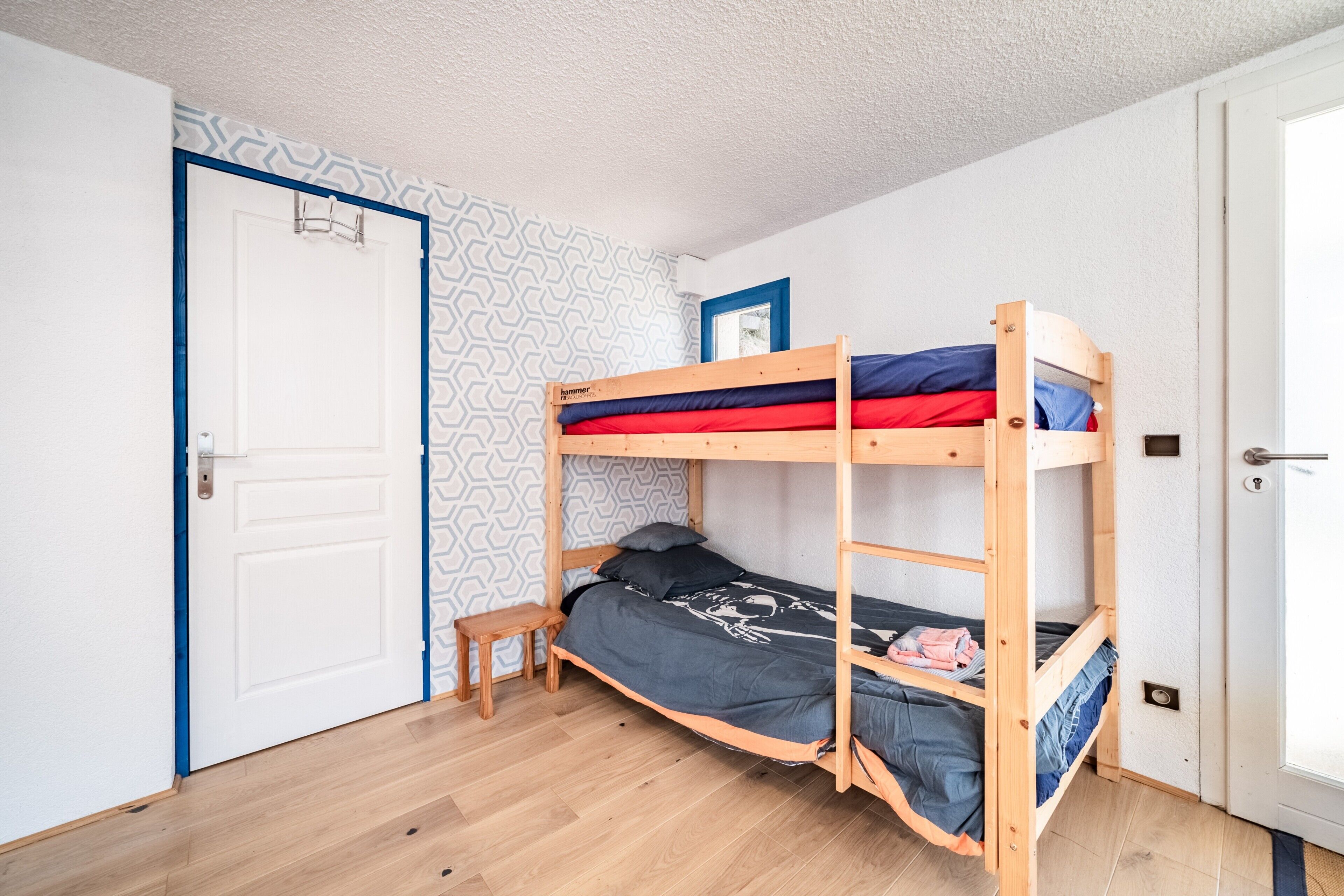 Une chambre avec un lit superposé, une porte, un petit tabouret et un papier peint à motifs.
