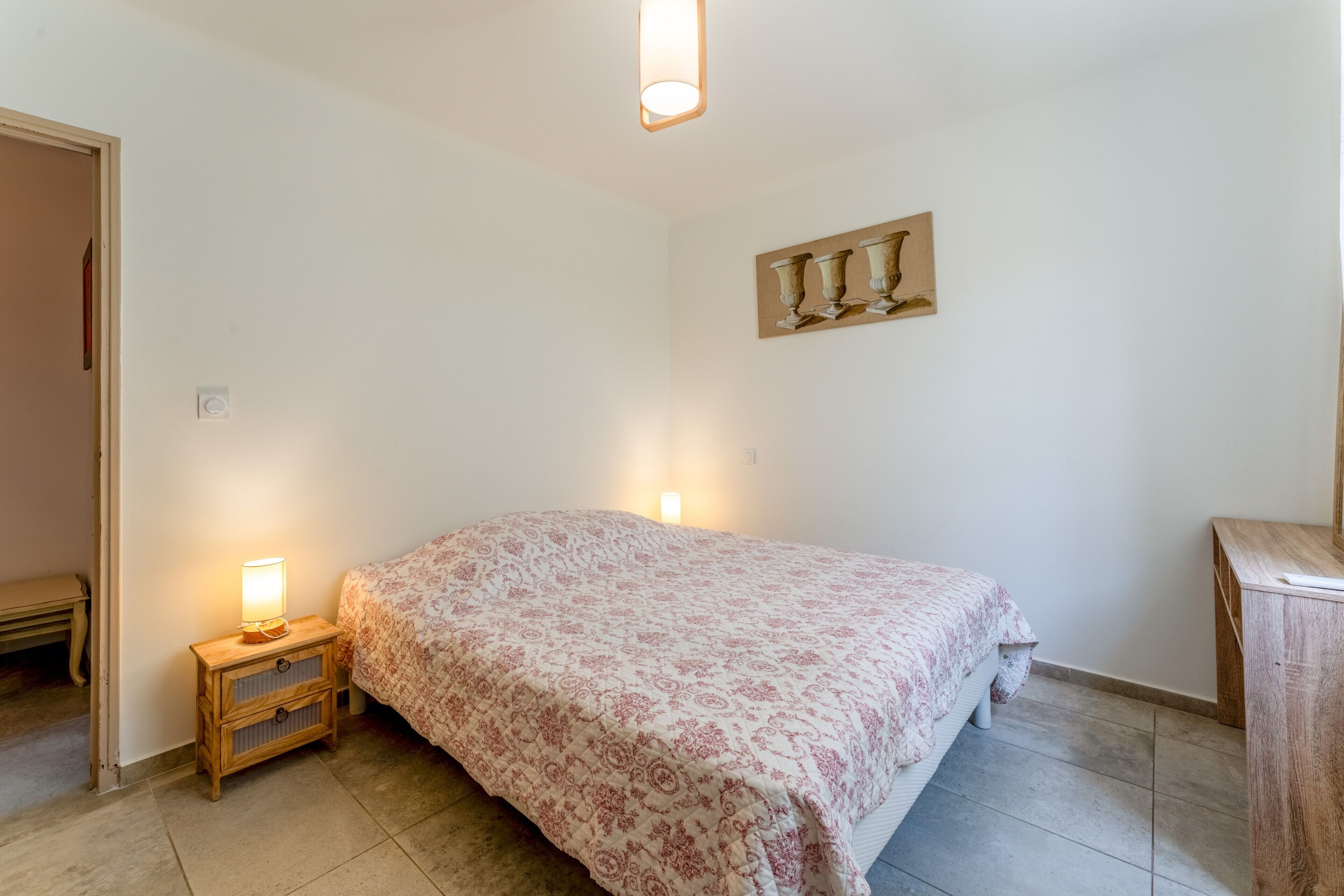A bedroom with a bed, bedside lamps, a wooden nightstand, a wall-mounted picture, and a door.