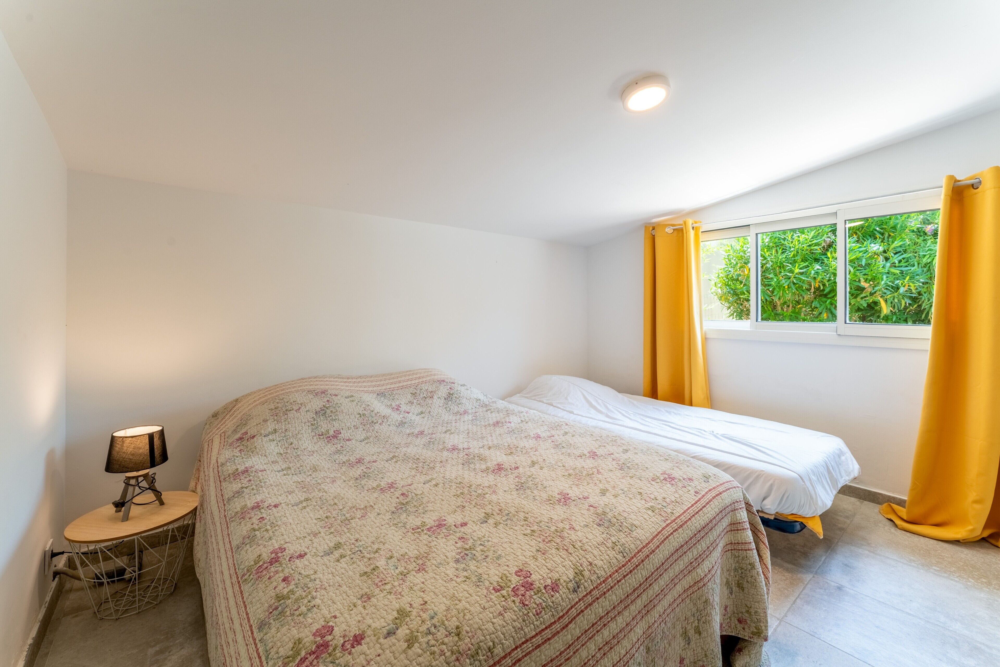 Ein Schlafzimmer mit Bett, Nachttisch, Lampe und einem Fenster mit Vorhängen.
