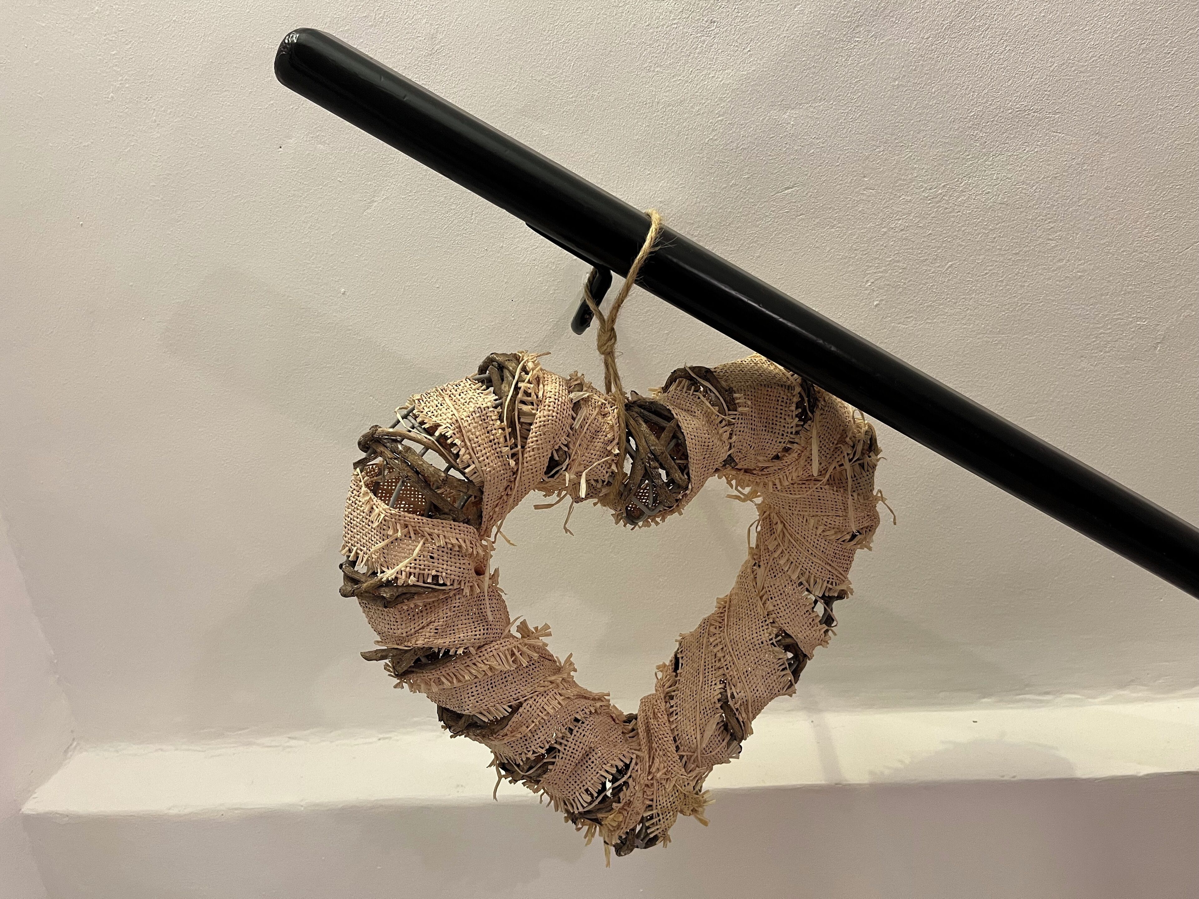 A heart-shaped decoration made of burlap and twine, hanging from a black rod against a white wall.
