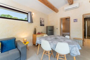 Een verzorgde kamer met een eettafel, stoelen, een bank, een klein bijzettafeltje met een lamp, een koelkast en een raam met uitzicht op groen.