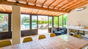 Un espacio de comedor al aire libre con techo, una mesa y sillas, una piscina al fondo y una zona de cocina con un lavamanos.