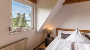 A bedroom with a sloped ceiling, a window with a view, a bedside table with a lamp, and a bed with pillows. - Apartment 2 in Haus Böhler on the island of Reichenau with Lake View, Terrace and Garden (Reichenau)