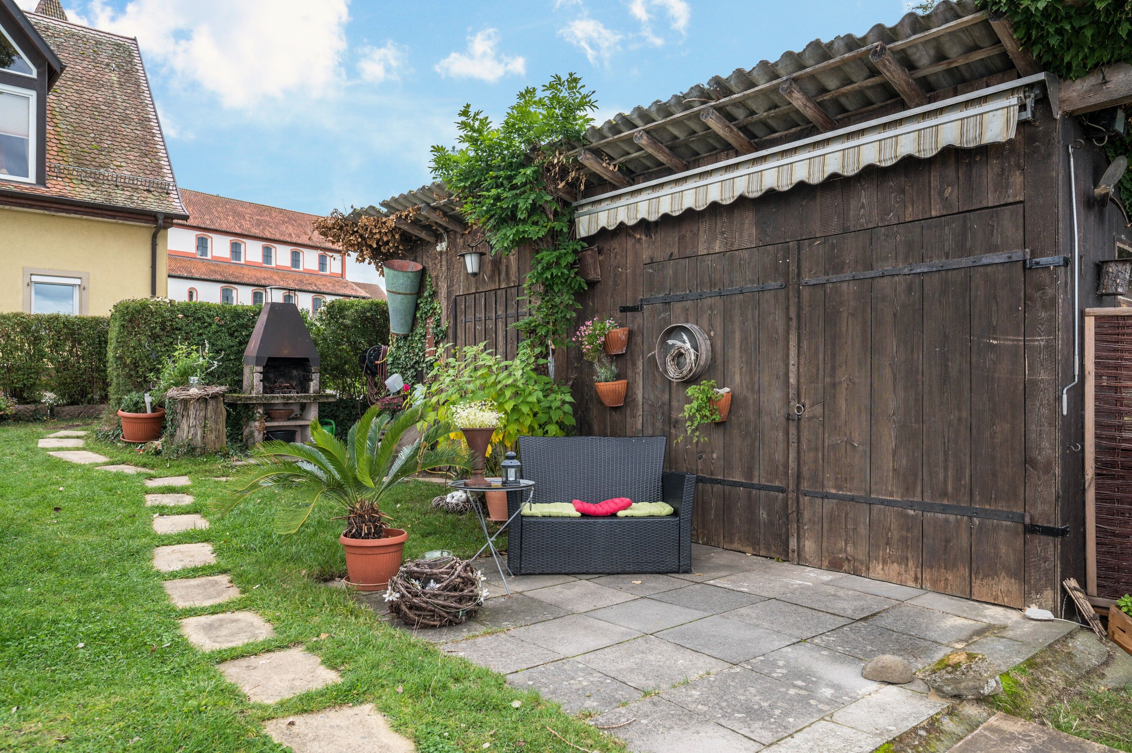 Un patio con una parrilla, plantas en macetas, una silla de mimbre y un portón de madera.