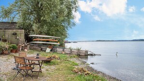 Une structure en bois au bord du lac, avec une table et des chaises, entourée de plantes en pot et d’une rangée de canoës.