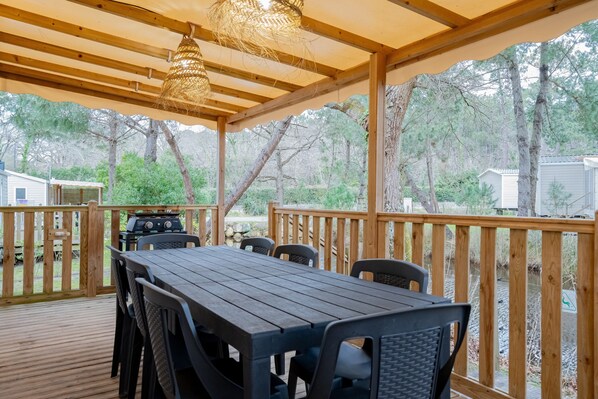 A covered outdoor deck with a table and chairs, a grill, and a view of a wooded area. - The Bassin Cabin – 3-Bedroom Mobile Home with Terrace and Pool (Lège-Cap-Ferret)