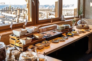 Disposição de buffet com diversos alimentos, utensílios, mobiliário de madeira e vista para a cidade através de grandes janelas.