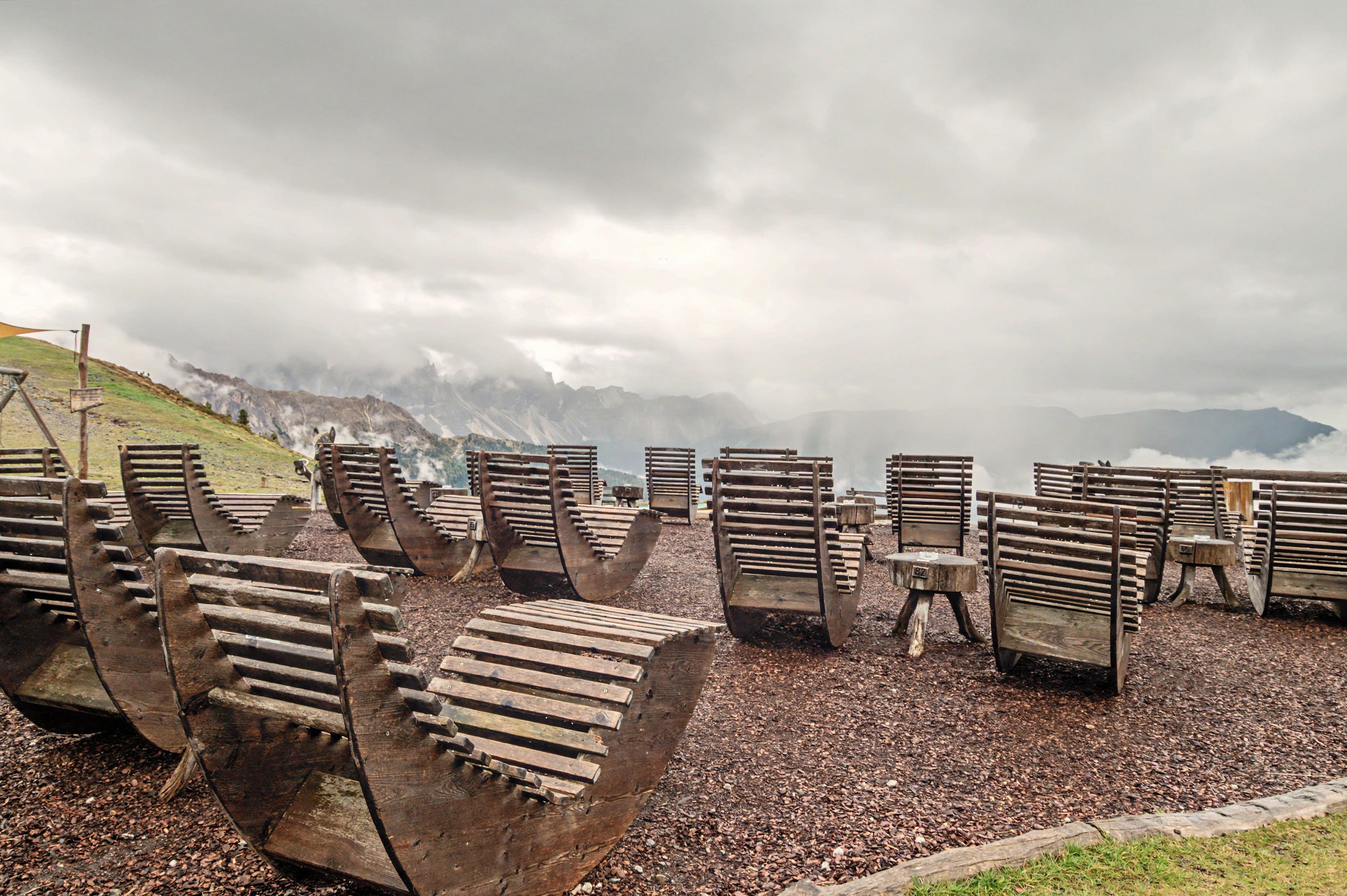A collection of rustic wooden chairs arranged on a gravel surface with a mountainous backdrop.