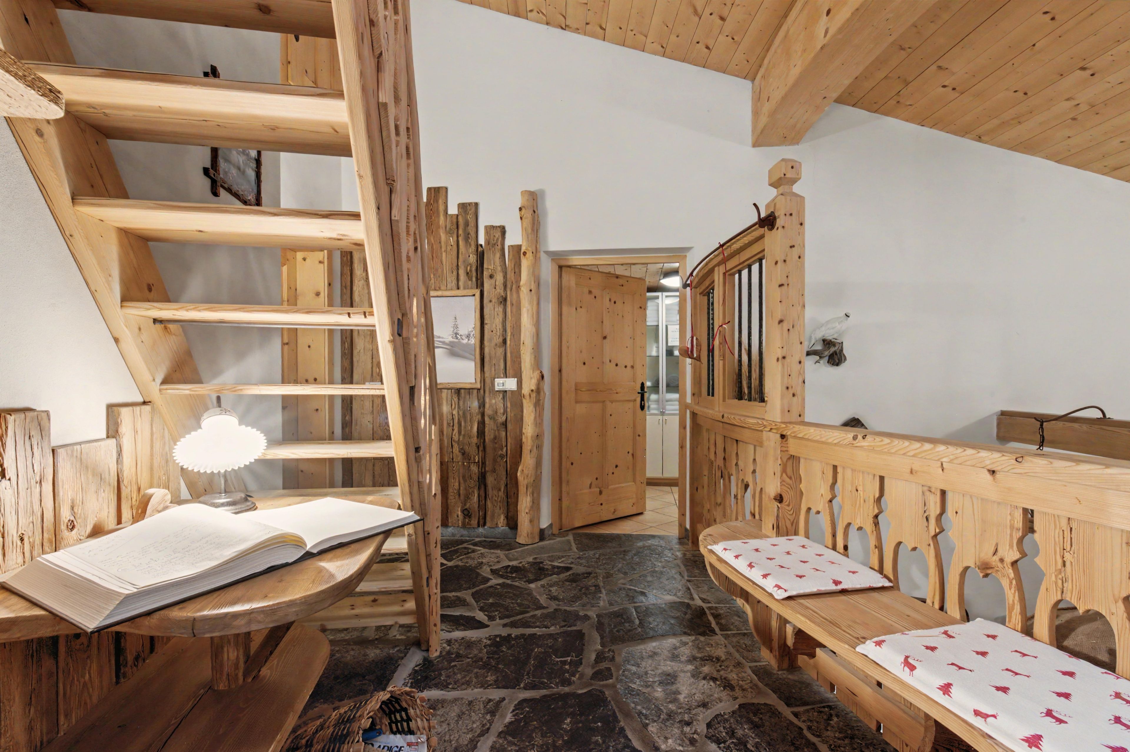 Un interno rustico in legno con pavimento in pietra, una panca di legno, un tavolo con un libro e una scala.