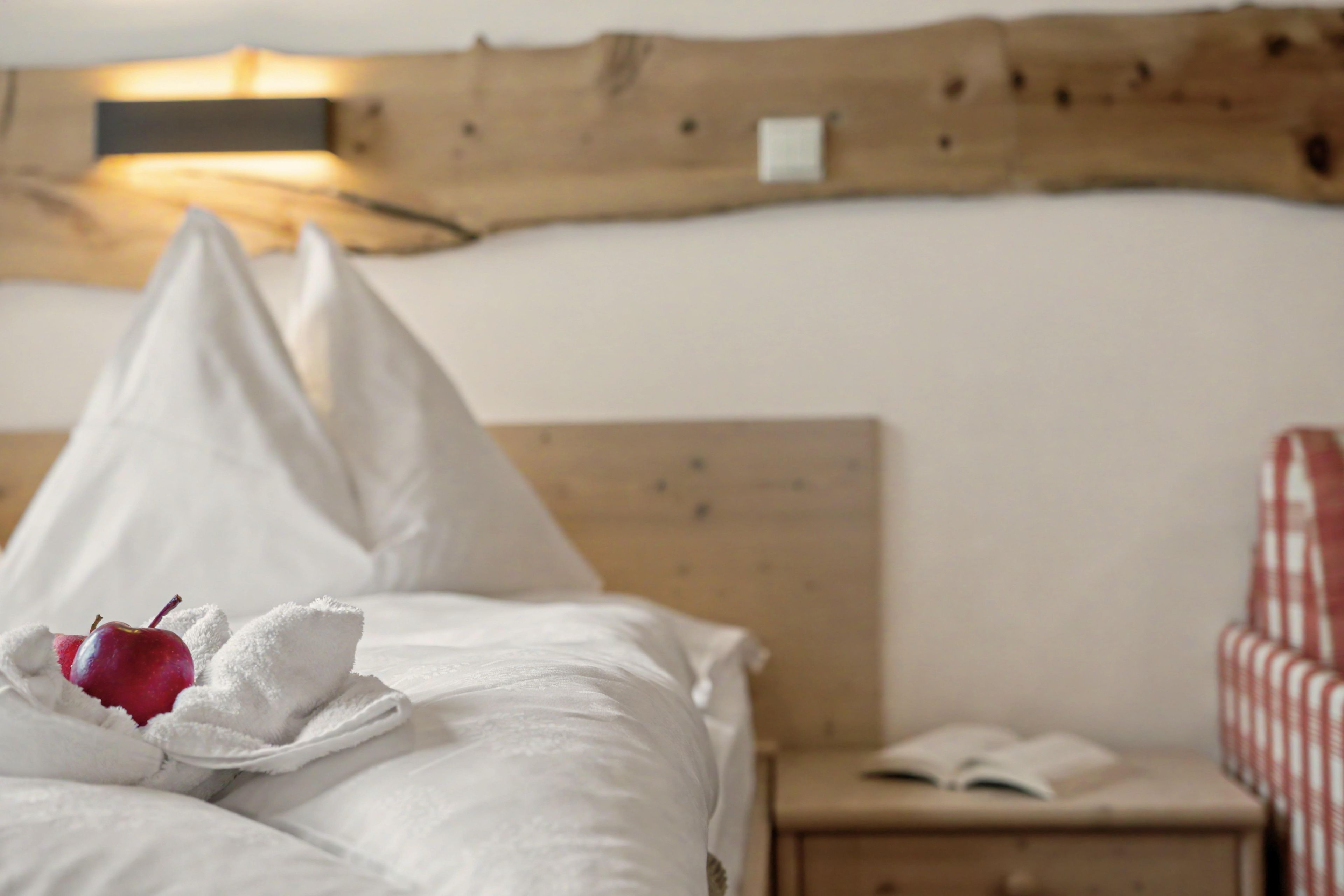 Un letto con lenzuola bianche, una mela rossa e un asciugamano. Si nota un poggiatesta in legno e un comodino con un libro aperto.