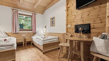 Habitación tipo cabaña de madera con una cama, dos mesitas de noche, una pequeña mesa con una silla, un televisor montado en la pared y una ventana con cortinas.