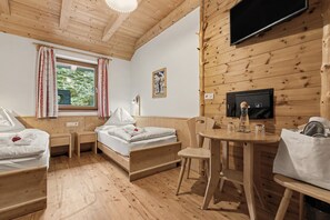 A wooden cabin room with a bed, two bedside tables, a small table with a chair, a TV mounted on the wall, and a window with curtains.