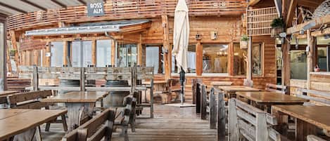 Un intérieur rustique en bois, avec de nombreuses tables et bancs en bois, une enseigne indiquant « ROSS ALM » et un espace extérieur abrité avec des sièges.