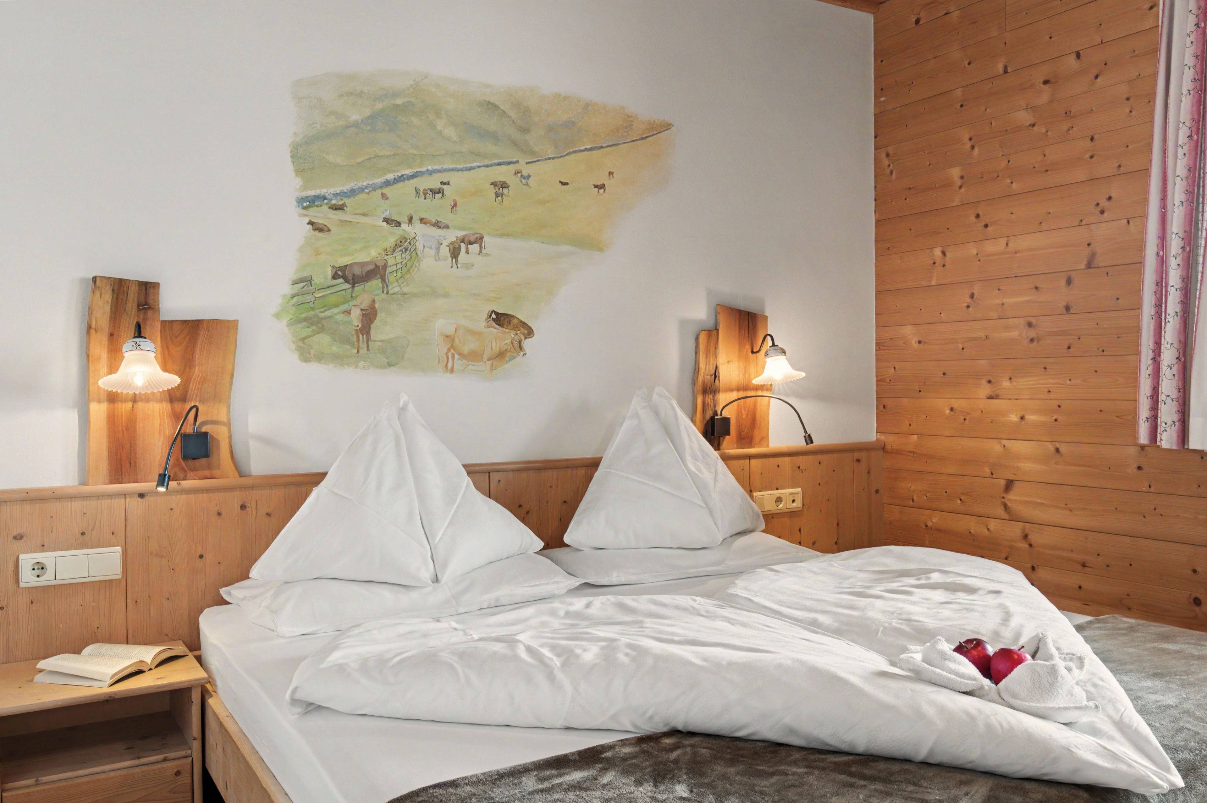 Quarto com cama, duas mesas de cabeceira, luminária na parede e pintura de vacas em uma fazenda.