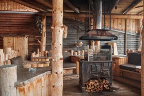 A rustic wooden interior with a large wood-burning stove, benches, and a glass-top table. - Room 'Rossalm Panoramic Room 4' with Shared Terrace, Shared Garden and Wi-Fi (Brixen)