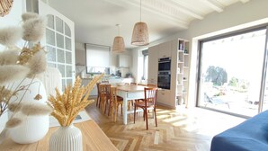 A modern kitchen with white cabinetry, a dining area with wooden chairs, and a large window offering a view of the outdoors. - La Perle, 6 guests, 180° sea view, jacuzzi (Plobannalec-Lesconil)