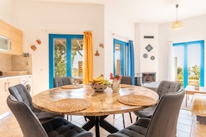 A dining area with a round wooden table, grey upholstered chairs, and a fruit bowl centerpiece. - Holiday Home 'Kalliope' with Sea View, Wi-Fi and Air Conditioning (Gennadi - Rhodos - Dodekanese)