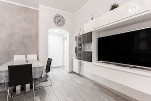 Un salon moderne doté d’un téléviseur à écran plat, d’un coin repas avec une table et des chaises, et d’une cuisine équipée d’un réfrigérateur et d’une horloge murale.