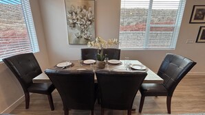 Uma sala de jantar com uma mesa retangular, quatro cadeiras pretas e um vaso com flores brancas. Há uma pintura na parede acima da mesa.