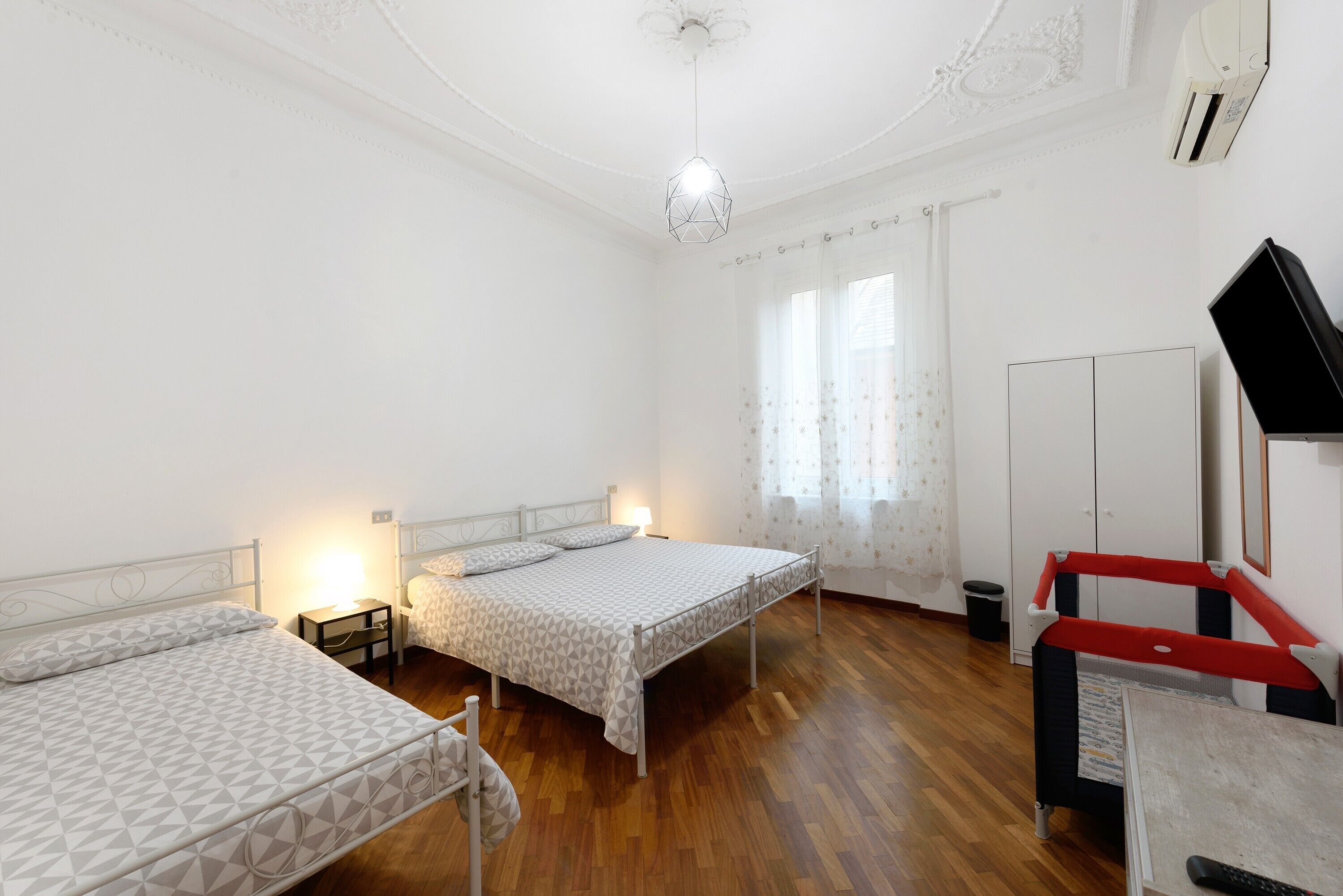 A room with two beds, a desk, a chair, a ceiling light, and a window with curtains.