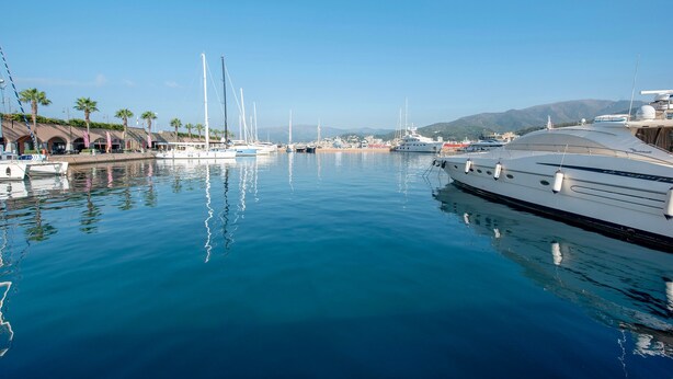 A marina with boats docked, calm water, and a mountainous backdrop.