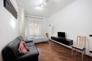 A modern living room with a black leather sofa, a wooden chair, a TV on a black stand, and a bed with a white bedspread. - Apartment 'Leo Genova Aeroporto' with Wi-Fi (Genova)