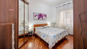 A bedroom with a wooden wardrobe, a bed with floral bedding, a nightstand, a wall-mounted painting, and a window with sheer curtains.