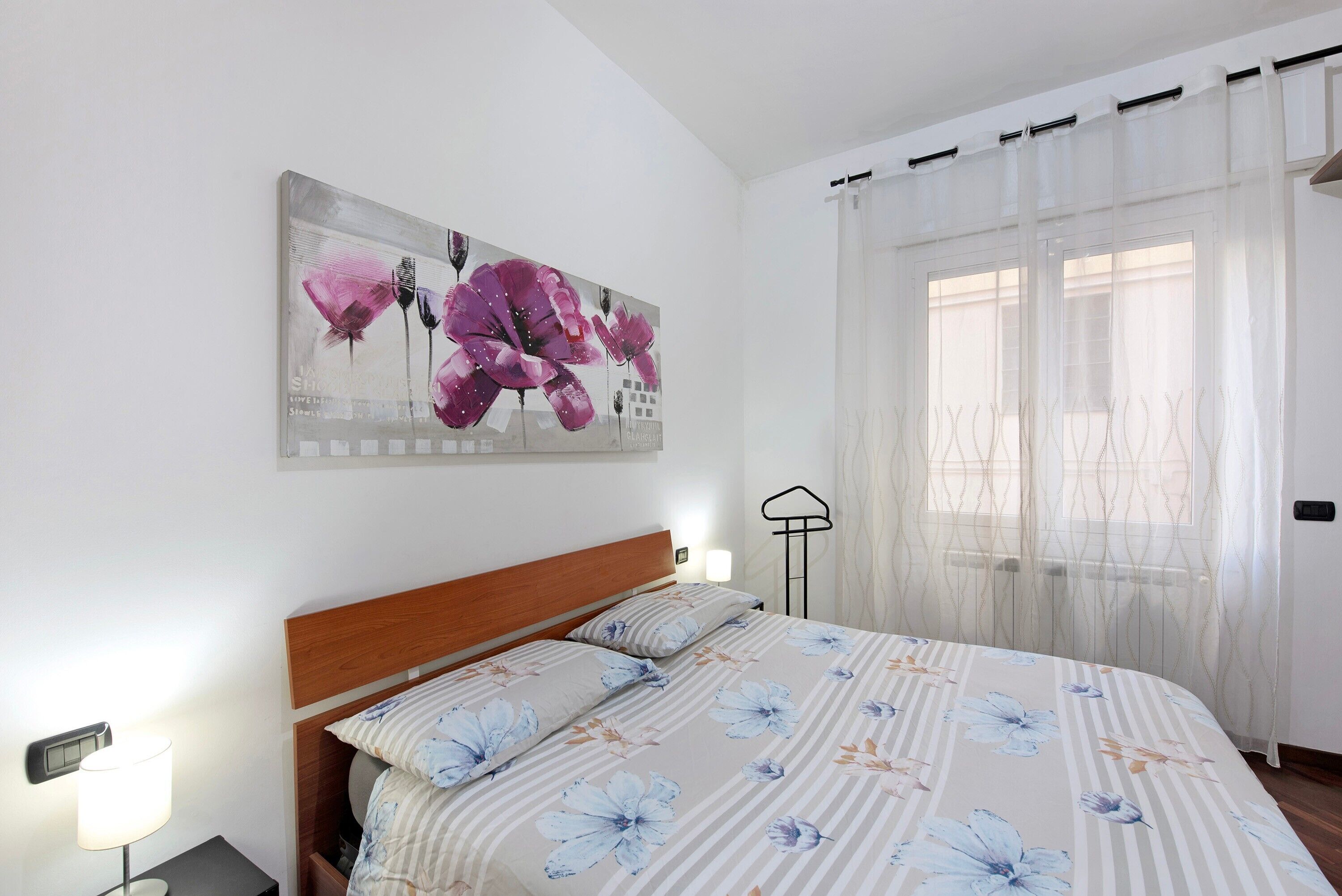 A bedroom with a bed, a nightstand, a wall-mounted lamp, a painting, and a window with sheer curtains.
