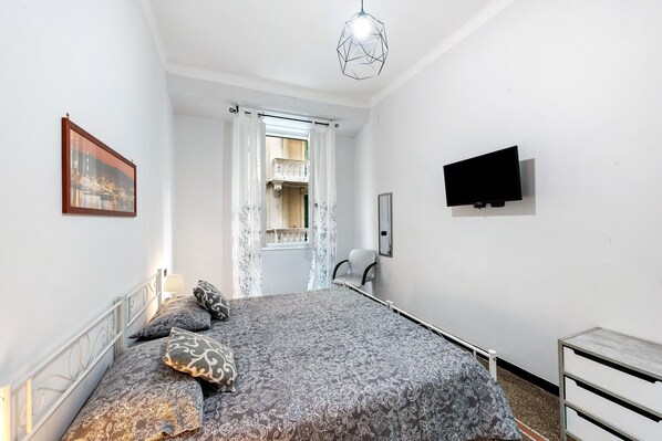 Un dormitorio con cama, televisor, silla, ventana con cortinas e imagen en la pared.