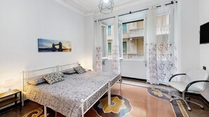 Un dormitorio con cama, una silla, un ventanal con cortinas, un cuadro y una alfombra estampada.