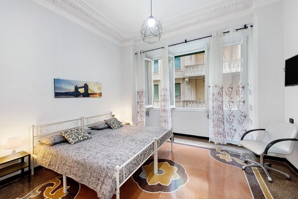 A bedroom with a bed, a chair, a window with curtains, a painting, and a patterned rug.