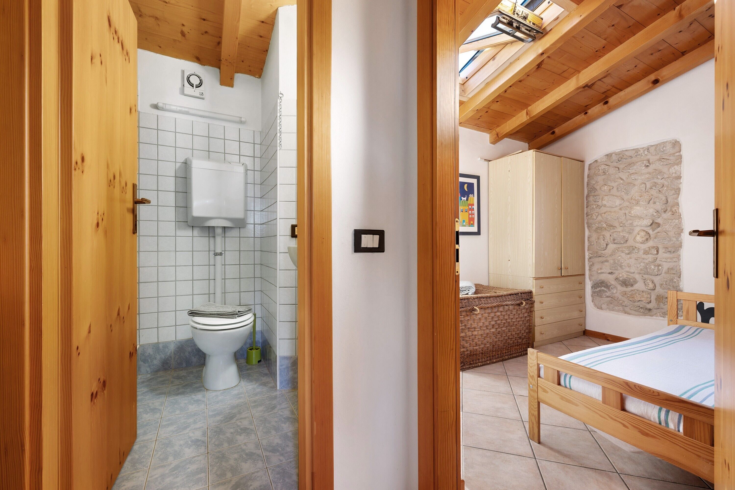 A wooden-framed room with a bed, a stone wall, and a tiled bathroom with a toilet and a towel rack.