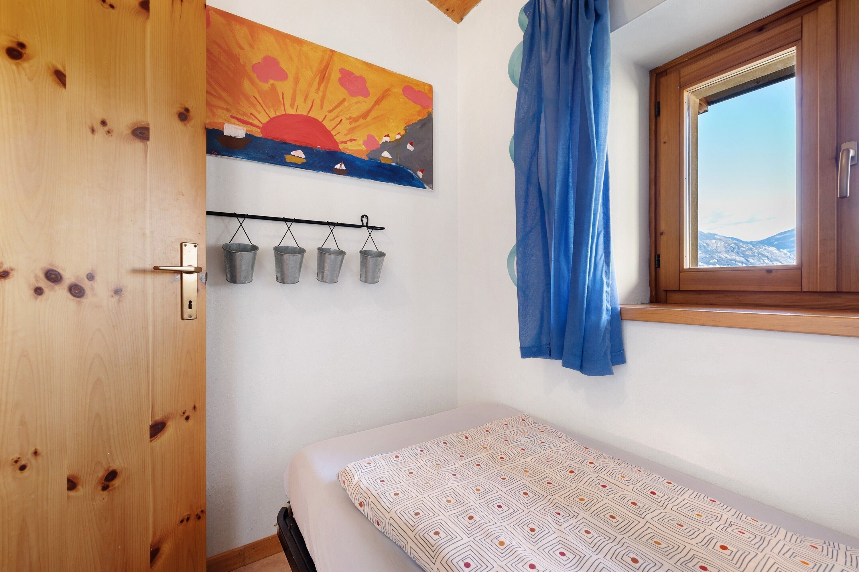 Une chambre à coucher avec un lit, une porte en bois, une fenêtre donnant sur un paysage, un tableau et un présentoir en métal avec des seaux.