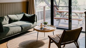 Ein modernes Wohnzimmer mit einer Couch, einem runden Couchtisch und einem Sessel. Ein großes Fenster bietet Blick auf eine Terrasse.