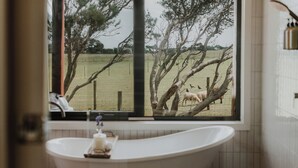 Ein Badezimmer mit freistehender Badewanne, ein Fenster mit Blick auf einen umzäunten Außenbereich mit Bäumen und Tieren.