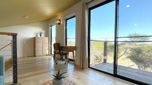 A spacious room with large windows, a wooden floor, a desk, a chair, and a small table with a plant.
