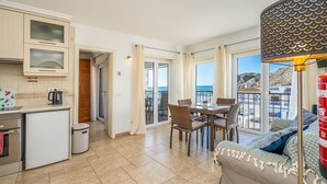 A kitchen with a refrigerator, stove, and cabinets, a dining area with chairs and a table, and a balcony with a view of the sea.